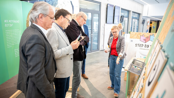 Eine Mit-Organisatorin einer Demokratie-Ausstellung erkl&auml;rt mehreren Besuchern eine interaktive Ausstellungsstation. Die Frau steht rechts und zeigt auf ein Exponat, w&auml;hrend drei M&auml;nner aufmerksam zuh&ouml;ren und Infomaterial in den H&auml;nden halten. Die Szene spielt in einem hellen Ausstellungsraum mit Infotafeln zum Thema Demokratie.