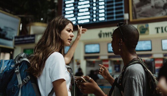 Zwei junge Frauen mit Reiserucks&auml;cken stehen in einem Bahnhof oder Flughafen und sprechen miteinander. Eine h&auml;lt ein Smartphone in der Hand, die andere gestikuliert erkl&auml;rend. Im Hintergrund ist eine gro&szlig;e Abflugtafel sowie ein Ticketschalter zu sehen.
