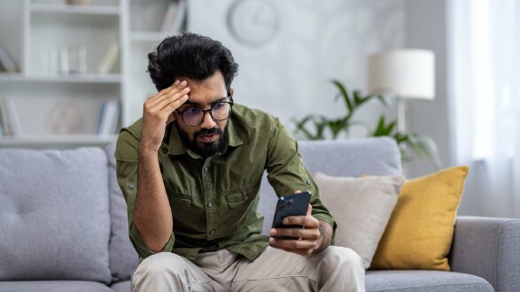 Ein Mann sitzt auf dem Sofa und schaut verzweifelt auf sein Smartphone.