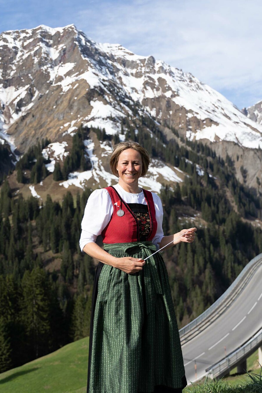 Angela Schwarzmann steht in Tracht und mit einem Takstock in der Hand vor einem beeindruckenden Bergpanorama und lächelt in die Kamera.