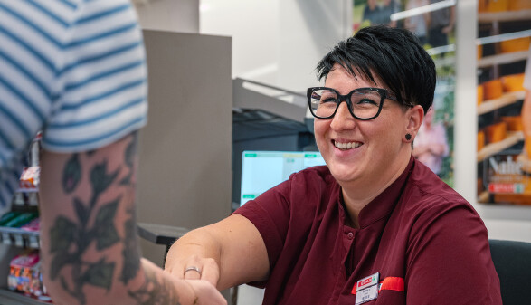 Eine l&auml;chelnde Kassiererin mit Brille, kurzen dunklen Haaren und einem weinroten SPAR-Arbeitshemd sitzt an einer Kasse. Sie reicht einer anderen Person, deren t&auml;towierter Arm und gestreiftes Oberteil im Vordergrund zu sehen sind, Wechselgeld oder ein Objekt.