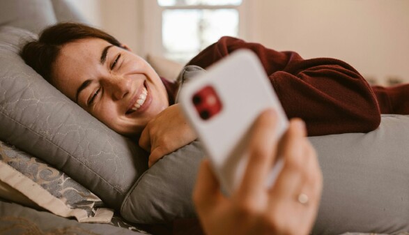 Eine Frau liegt lächelnd im Bett und hält ein Handy in der Hand. 