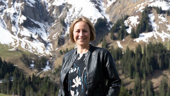 Eine Frau mit halblangen, blonden Haaren, Angela Schwarzmann, lächelt in die Kamera. Im Hintergrund sieht man ein beeindruckendes Bergpanorama, auf dem teils noch Schnee liegt.