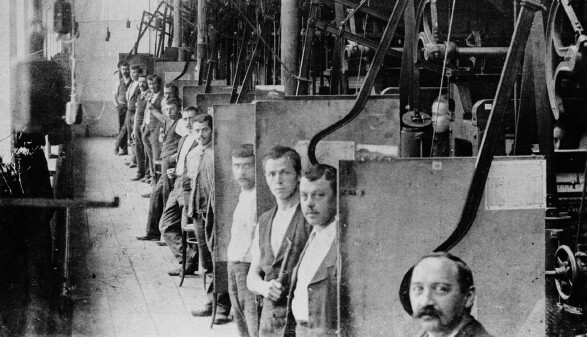 Eine historische Schwarz-Wei&szlig;-Aufnahme aus dem Inneren einer Fabrik. Eine lange Reihe von Arbeitern steht hintereinander an ihren jeweiligen Arbeitsstationen, die durch trennende Platten voneinander abgeteilt sind. Die M&auml;nner blicken alle in Richtung der Kamera. Im Vordergrund ist das Gesicht eines Mannes mit Schnurrbart angeschnitten zu sehen. Entlang der Decke und W&auml;nde verlaufen komplexe mechanische Vorrichtungen, Riemen und Maschinenteile. Der Boden besteht aus Holzdielen.