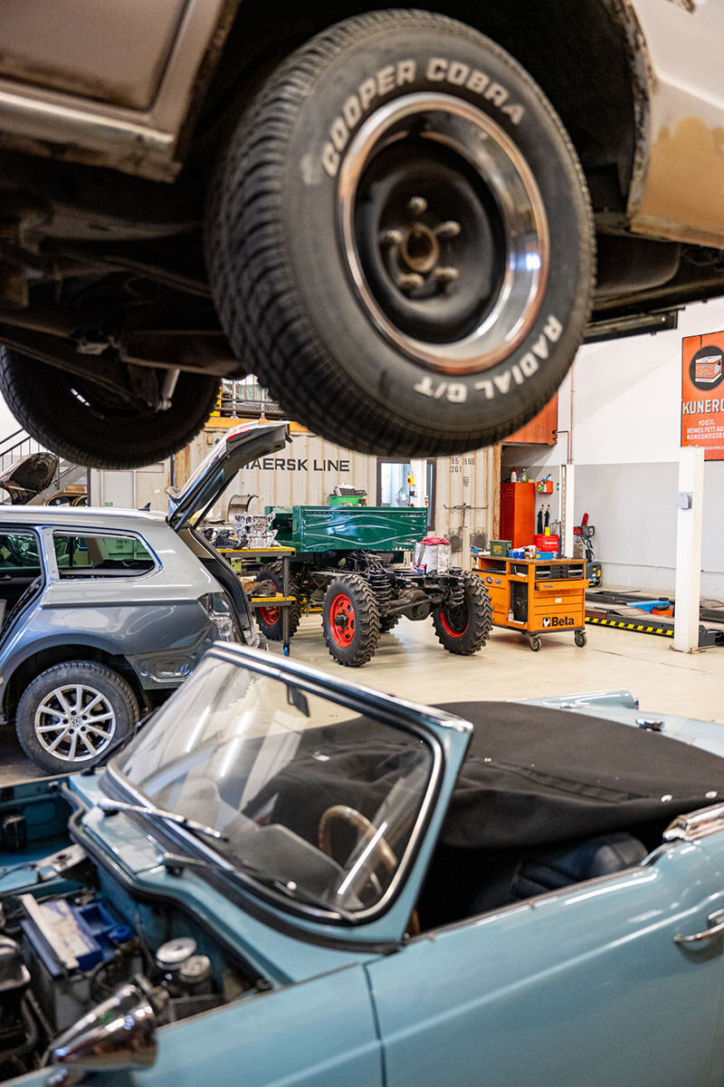 Blick in Werner Metzlers Werkstatt: Im Vordergrund ein hellblauer Oldtimer-Cabriolet mit ge&ouml;ffneter Motorhaube, dar&uuml;ber ein auf der Hebeb&uuml;hne angehobenes Fahrzeug mit Cooper-Cobra-Reifen. Im Hintergrund stehen weitere Fahrzeuge und Fahrzeugrahmen in der ger&auml;umigen Halle.