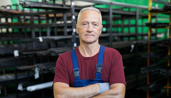 &Auml;lterer Arbeiter in Latzhose steht mit verschr&auml;nkten Armen in einem Industrielager und blickt ernst in die Kamera.