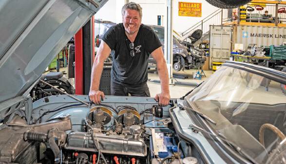 Werner Metzler l&auml;chelt in die Kamera, w&auml;hrend er sich &uuml;ber den ge&ouml;ffneten Motorraum eines Oldtimers beugt. Im Hintergrund ist eine gut ausgestattete Werkstatt zu sehen.