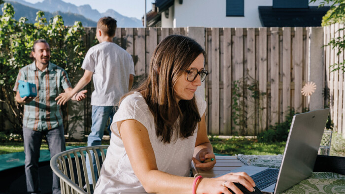 Eine Frau mit Brille sitzt an einem Tisch im Garten und arbeitet an einem Laptop. Im Hintergrund stehen ein Mann und ein Jugendlicher auf dem Rasen vor einem Holzzaun und unterhalten sich. Im Hintergrund erstreckt sich eine Bergkulisse.