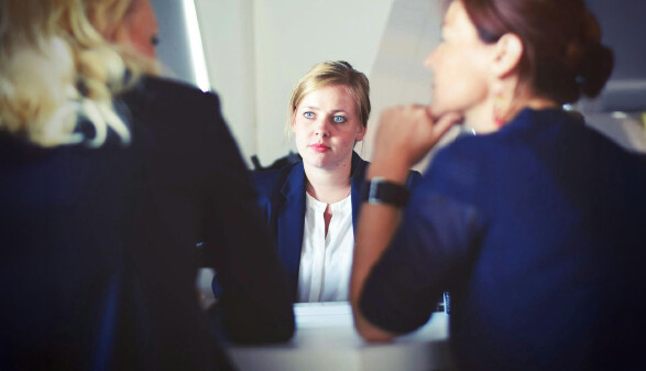 Eine verunsichert und nervös aussehende blonde Frau sitzt bei einem Bewerbungsgespräch zwei Frauen gegenüber, die miteinander reden.