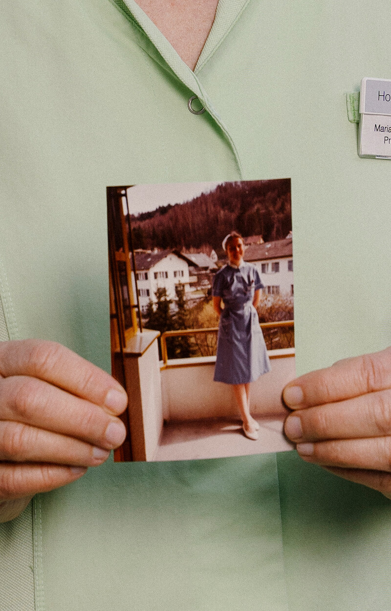 Maria Lackner h&auml;lt ein altes Foto aus den 1980er-Jahren in den H&auml;nden, das sie als junge Krankenschwester in hellblauer Uniform und wei&szlig;er Haube auf einem Balkon zeigt. Im Hintergrund des historischen Fotos sind Geb&auml;ude und ein bewaldeter Hang zu sehen. Maria Lackner tr&auml;gt heute ihren hellgr&uuml;nen Pflegekittel mit Namensschild.