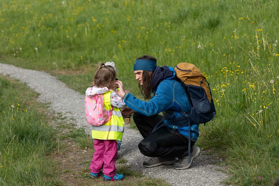 Ein Mann mit langen, braunen Haaren und einem großen Rucksack auf dem Rücken, Simon Engstler, kniet vor einem kleinen Mädchen und hilft ihr, eine Haarsträhne aus dem Gesicht zu streichen.