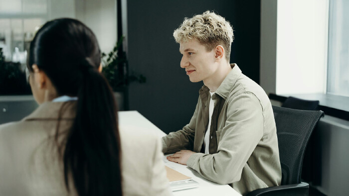 Ein junger, blonder Mann in einem Hemd sitzt in einer Gesprächssituation mit einer dunkelhaarigen Frau in einem Blazer an einem Besprechungstisch und lächelt selbstsicher.