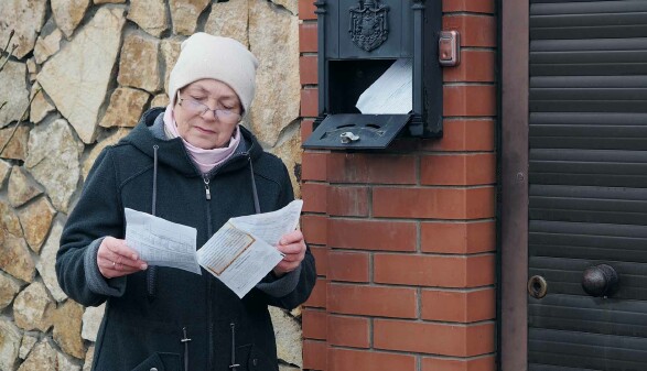 Eine ältere Frau steht vor dem Briefkasten und liest einen Brief.