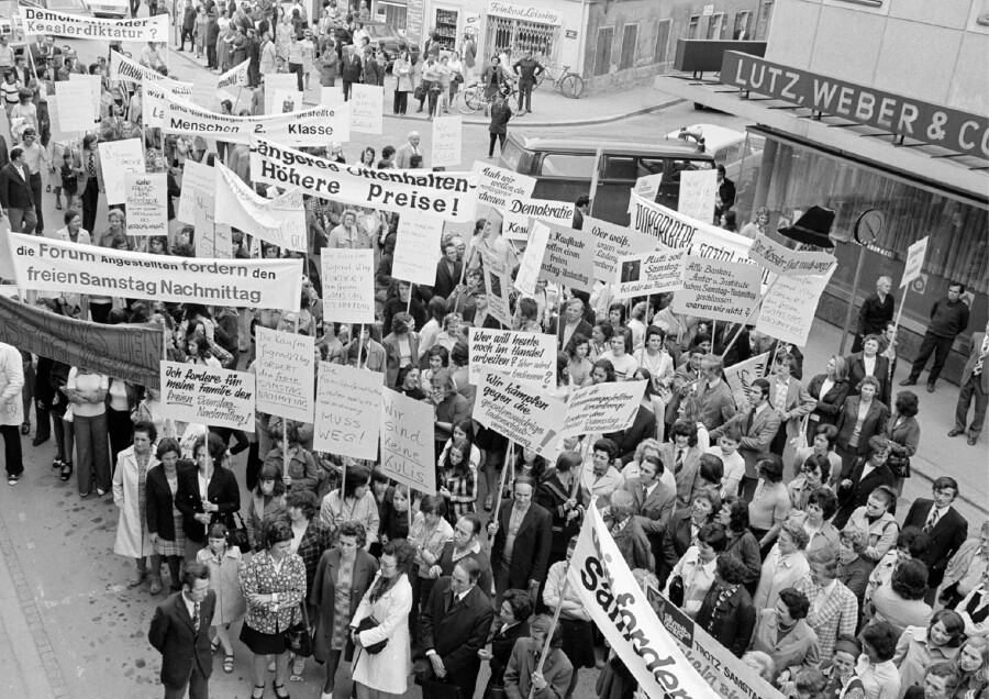 Schwarz-Wei&szlig;-Vogelperspektive einer gro&szlig;en Demonstration in einer Stadtstra&szlig;e. Zahlreiche Menschen ziehen mit Schildern und Transparenten durch die Stra&szlig;e. Auf den Transparenten stehen Forderungen wie &bdquo;die Forum Angestellten fordern den freien Samstag Nachmittag&ldquo;, &bdquo;L&auml;ngeres Offenhalten &ndash; H&ouml;here Preise!&ldquo; und &bdquo;Demokratie oder Ke&szlig;lerdiktatur?&ldquo;. Rechts sind Gesch&auml;fte wie &bdquo;Feinkost Leissing&ldquo; und &bdquo;LUTZ, WEBER & CO&ldquo; zu sehen.