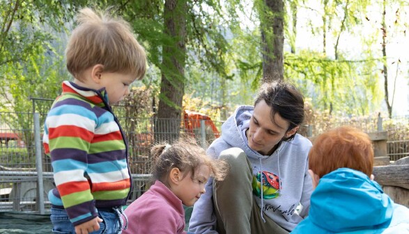 Ein Mann mit langen, braunen Haaren, Simon Engstler, kniet in einem Sandkasten. Um ihn herum stehen drei kleine Kinder, ein kleines Mädchen spielt mit ihm im Sand.