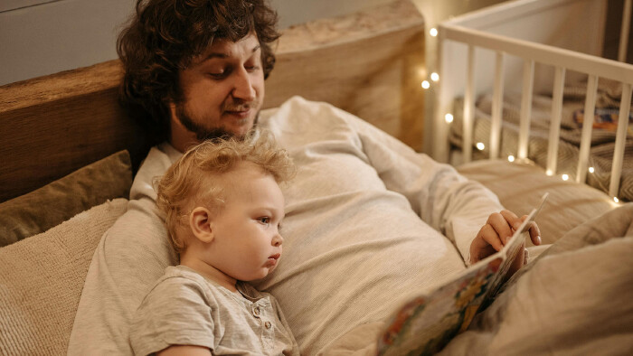 Ein Vater liegt mit seinem Kleinkind im Bett und liest ihm ein Bilderbuch vor. Im Hintergrund sind ein wei&szlig;es Kinderbett und kleine Lichterketten zu sehen. Das Bild steht im Kontext einer Familie, die von einer gro&szlig;z&uuml;gigen Steuergutschrift profitiert.