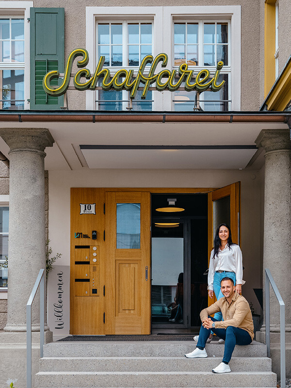 Au&szlig;enansicht des Eingangs der Schaffarei Gastronomie. &Uuml;ber dem Vordach ist ein gro&szlig;er, gelber Neonschriftzug mit dem Namen &bdquo;Schaffarei&ldquo; angebracht. Auf den Steinstufen des &uuml;berdachten Eingangs, der von zwei grauen S&auml;ulen flankiert wird, befinden sich die P&auml;chter: Adrien sitzt im Vordergrund auf den Stufen, Alyssa steht leicht versetzt dahinter neben der offenen Holzt&uuml;r. Links neben der T&uuml;r h&auml;ngt ein Schild mit der Aufschrift &bdquo;Willkommen&ldquo; und der Hausnummer 10.