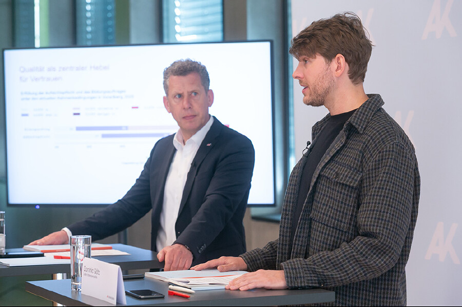 Pressegespr&auml;ch der Arbeiterkammer Vorarlberg. Dominic G&ouml;tz, &Ouml;konom der AK Vorarlberg, spricht stehend an einem Tisch, auf dem Unterlagen, Wassergl&auml;ser und ein Namensschild mit seinem Namen liegen. Er tr&auml;gt ein schwarzes T-Shirt unter einem grauen Karojackett und ein Ansteckmikrofon. Im Hintergrund sitzt AK-Pr&auml;sident Bernhard Heinzle im dunklen Sakko mit wei&szlig;em Hemd. Hinter den beiden ist eine Pr&auml;sentationsfolie mit der &Uuml;berschrift &bdquo;Qualit&auml;t als zentraler Hebel f&uuml;r Vertrauen" zu sehen. Foto: Dietmar Mathis.