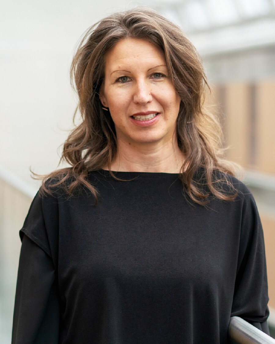 Portr&auml;t einer l&auml;chelnden Frau mit mittellangen, leicht gewellten braunen Haaren und schwarzem Oberteil. Sie blickt direkt in die Kamera. Der Hintergrund ist hell und verschwommen.
