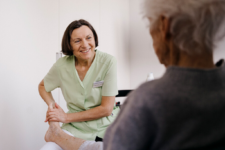 Krankenschwester Maria Lackner l&auml;chelt eine &auml;ltere Patientin an, w&auml;hrend sie deren Fu&szlig; bzw. Unterschenkel behutsam in den H&auml;nden h&auml;lt. Maria Lackner tr&auml;gt einen hellgr&uuml;nen Pflegekittel mit Namensschild. Die Patientin ist von hinten zu sehen und sitzt ihr gegen&uuml;ber.