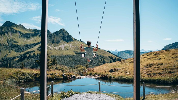 Das Foto zeigt eine Frau auf einer Schaukel. Direkt vor ihr liegt ein schöner, blauer Bergsee, im Hintergrund sieht man Berggipfel, grüne Wiesen und blauen Himmel.