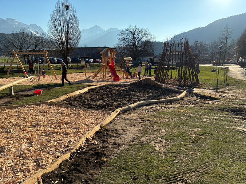Das Foto zeigt einen Spielplatz mit Sandkasten, Schaukel und Rutsche. Im Hintergrund sieht man mehrere Berggipfel.