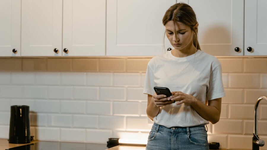 Eine Frau steht vor einem Küchenschrank und schaut auf ein Handy in ihrer Hand.