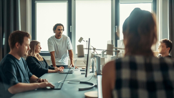 Fünf Personen in einem hellen Büro sitzen und stehen an einem langen Tisch, arbeiten an Laptops und diskutieren in entspannter Atmosphäre.