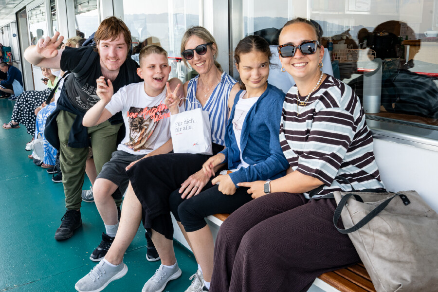 Eine Gruppe von Fahrg&auml;sten sitzt gut gelaunt nebeneinander auf einer Holzbank an Deck eines Passagierschiffs. Ganz rechts sitzt eine l&auml;chelnde Frau mit Sonnenbrille und einem braun-wei&szlig; gestreiften T-Shirt, neben ihr eine junge Frau in einer blauen Strickjacke. In der Mitte h&auml;lt eine Frau mit Sonnenbrille und gestreifter Bluse eine wei&szlig;e Papiert&uuml;te mit der Aufschrift &bdquo;F&uuml;r dich, f&uuml;r alle. AK Vorarlberg&ldquo;. Links von ihr zeigt ein Junge im wei&szlig;en T-Shirt fr&ouml;hlich das Peace-Zeichen, w&auml;hrend ein weiterer Jugendlicher im schwarzen T-Shirt im Hintergrund in die Kamera winkt. Alle wirken ausgelassen und freuen sich &uuml;ber den gemeinsamen Ausflug auf dem Wasser.