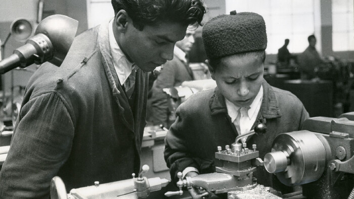 Schwarz-wei&szlig;-Bild aus dem Jahr 1960: Ausbildung eines jungen Lehrlings aus Algerien an einer Maschine zur Metallbearbeitung, ein anderer junger Mann erkl&auml;rt ihm und schaut ihm &uuml;ber die Schulter.