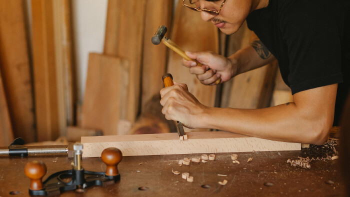 Das Foto zeigt einen jungen Mann in einer Werkstatt, der ein Holzwerkst&uuml;ck bearbeitet.