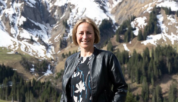 Eine Frau mit halblangen, blonden Haaren, Angela Schwarzmann, lächelt in die Kamera. Im Hintergrund sieht man ein beeindruckendes Bergpanorama, auf dem teils noch Schnee liegt.