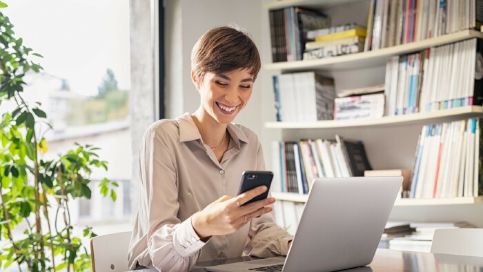 © francescoridolfi.com, stock.adobe.com Frau sitzt lachend am Schreibtisch mit einem Smartphone in der Hand. Vor ihr steht ein Notebook.