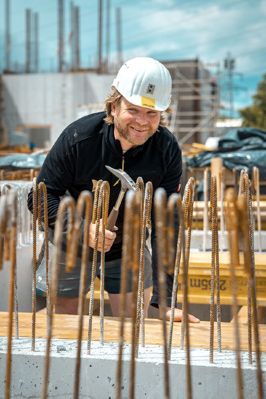 Ein Mann mit hellen Haaren und einem Bart, der als Hochbauer arbeitet, beugt sich l&auml;chelnd nach vorne und blickt direkt in die Kamera. Er tr&auml;gt eine schwarze Fleecejacke, kurze graue Arbeitshosen und einen wei&szlig;en Schutzhelm mit einem gelben Logo. In seiner rechten Hand h&auml;lt er einen Zimmermannshammer. Das Foto ist aus einer tiefen Perspektive aufgenommen, sodass im unmittelbaren Vordergrund eine Reihe von vertikalen, oben gebogenen Bewehrungsst&auml;hlen (Monatseisen) aus Beton herausragen, die den Blick leicht einrahmen. Er steht auf einer Baustelle; im Hintergrund sind weitere Baustrukturen, Betonw&auml;nde und ein bew&ouml;lkter blauer Himmel zu sehen.