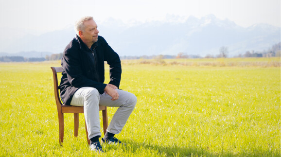 Oliver Loacker sitzt auf einem Stuhl auf einer Wiese, im Hintergrund ein Bergpanorama.