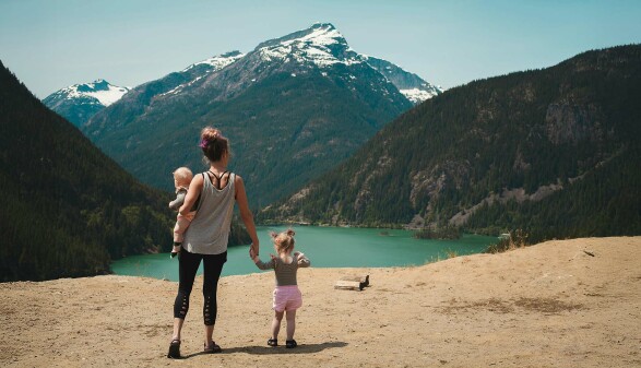 Eine Frau steht am Rande eines Sees. Auf dem Arm trägt sie ein Baby, an der anderen Hand hält sie ein kleines Mädchen. Im Hintergrund sieht man einen Berg mit schneebedecktem Gipfel.