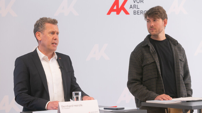 Pressegespr&auml;ch der Arbeiterkammer Vorarlberg. Zwei M&auml;nner stehen nebeneinander vor einer Stellwand mit dem Logo der AK Vorarlberg. Links spricht Bernhard Heinzle, AK-Pr&auml;sident &ndash; er tr&auml;gt ein dunkles Sakko und ein wei&szlig;es Hemd mit einem AK-Pin am Revers und ein Ansteckmikrofon. Vor ihm steht ein Namensschild mit der Aufschrift &bdquo;Bernhard Heinzle, AK Pr&auml;sident" sowie ein Wasserglas. Rechts steht AK &Ouml;konom Dominic G&ouml;tz in einem karierten Hemd &uuml;ber einem schwarzen T-Shirt, der aufmerksam zuh&ouml;rt.