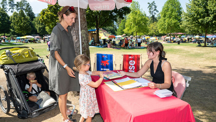 Eine Frau sitzt an einem rot gedeckten Stand der Arbeiterkammer im Freibad und &uuml;berreicht einem kleinen M&auml;dchen ein Buch, w&auml;hrend die Mutter danebensteht und ein Kleinkind im Kinderwagen sitzt.