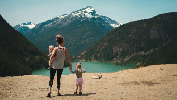 Eine Frau steht am Rande eines Sees. Auf dem Arm trägt sie ein Baby, an der anderen Hand hält sie ein kleines Mädchen. Im Hintergrund sieht man einen Berg mit schneebedecktem Gipfel.
