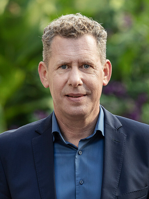 Portr&auml;t von Bernhard Heinzle mit kurzen, ergrauenden Kraushaaren, blauem Hemd und dunklem Sakko, der direkt in die Kamera blickt.