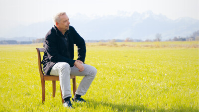 Oliver Loacker sitzt auf einem Stuhl auf einer Wiese, im Hintergrund ein Bergpanorama.