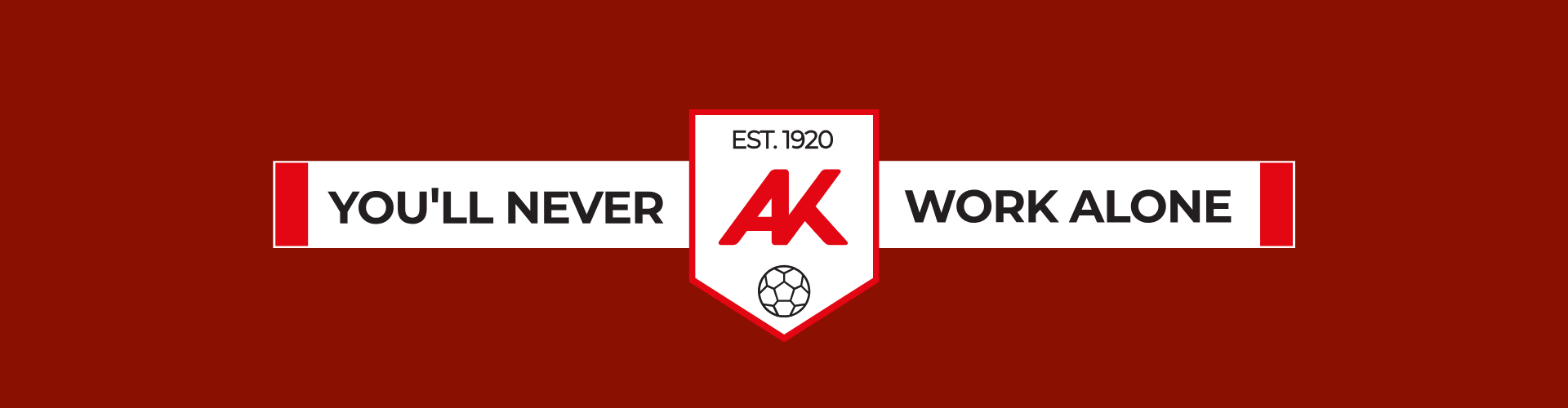 Ein dunkelrotes Banner mit einem zentralen wei&szlig;en Wappen, das die roten Buchstaben &bdquo;AK&ldquo;, das Gr&uuml;ndungsjahr &bdquo;EST. 1920&ldquo; und ein Fu&szlig;ball-Symbol enth&auml;lt. Links und rechts vom Wappen steht auf einem wei&szlig;en Balken in Gro&szlig;buchstaben der Slogan: &bdquo;YOU'LL NEVER WORK ALONE&ldquo;.