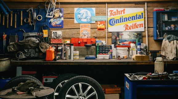 Eine detailreiche, rustikale Werkstatt mit einer h&ouml;lzernen R&uuml;ckwand, auf der verschiedene Vintage-Blechschilder (u.a. &bdquo;Continental Reifen&ldquo;, &bdquo;Osram&ldquo;, &bdquo;Vespa&ldquo;) h&auml;ngen. Eine lange Werkbank ist dicht mit Werkzeugen, Spraydosen, Kleinteilemagazinen und einem Kanister gef&uuml;llt. Im Vordergrund stehen Autoreifen auf Alufelgen.