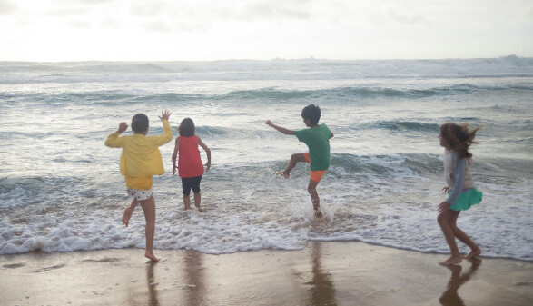 Vier Kinder spielen ausgelassen am Strand im Bereich der Brandung. Sie stehen mit dem R&uuml;cken zur Kamera und tragen bunte Sommerkleider in Gelb, Rot, Gr&uuml;n und Hellblau. Die Wellen rollen auf sie zu, eines der Kinder springt &uuml;ber das Wasser. Der Himmel ist leicht bew&ouml;lkt, das Meer rau.