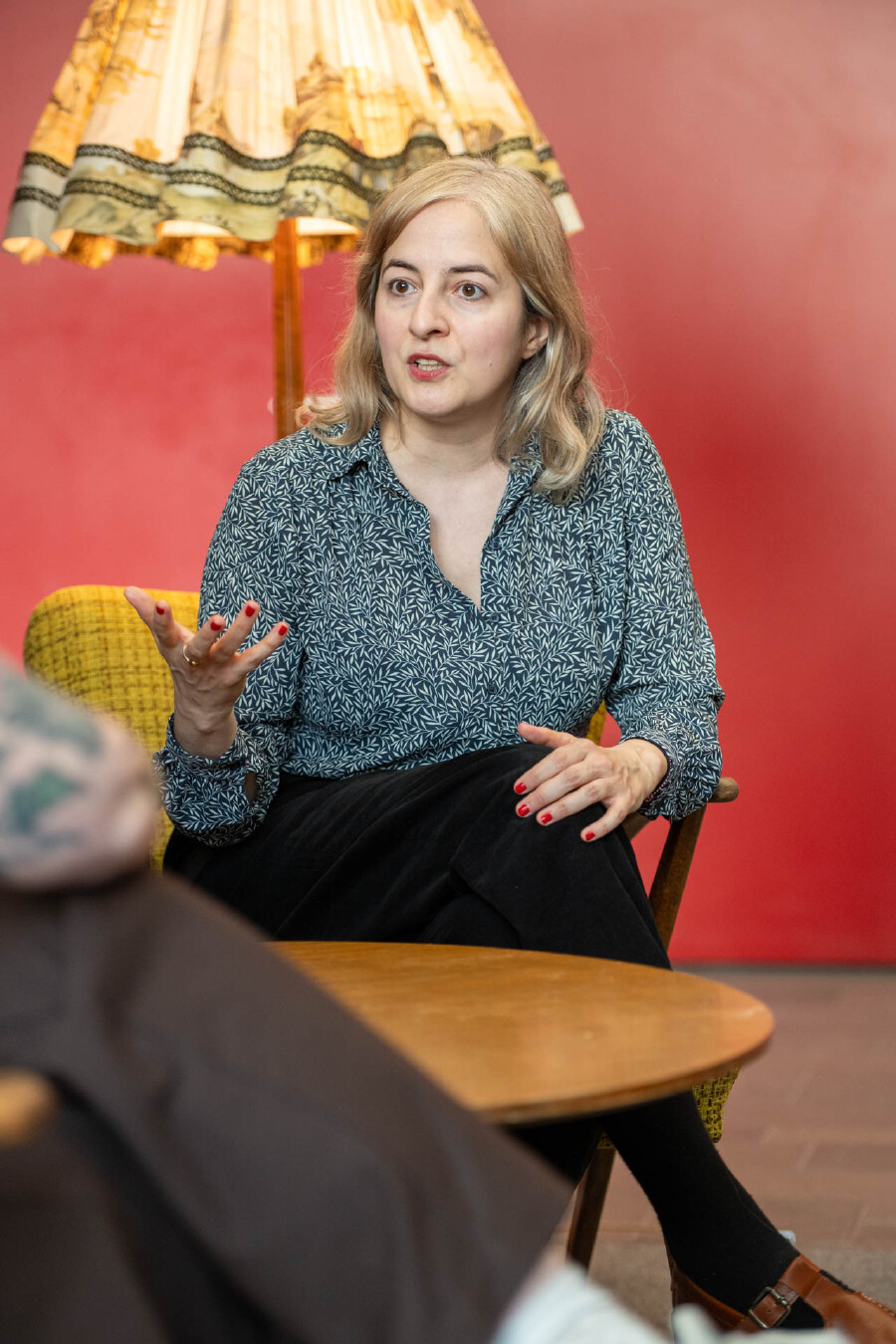 Eugenia Stamboliev sitzt auf einem gelben Sessel vor einer roten Wand und spricht mit einer erkl&auml;renden Handgeste zu ihrem Gegen&uuml;ber.