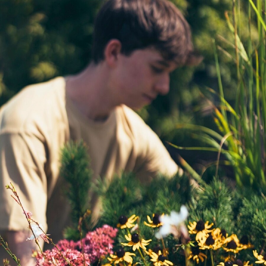 Gartenbau-Lehrling bei der Pflanzenpflege im blühenden Garten. Im Vordergrund farbenfrohe Blumen wie gelbe Margeriten und rosa Blüten, im Hintergrund eine Person in beigem Shirt bei der Arbeit. Symbolbild für praxisnahe Ausbildung im Garten- und Landschaftsbau, Frühling, Natur und nachhaltige Berufswahl.