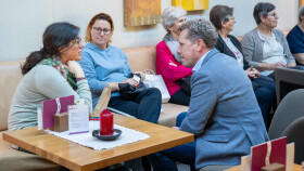 AK Pr&auml;sident Bernhard Heinzle sitzt rechts im Bild und beugt sich im Gespr&auml;ch zu zwei Teilnehmerinnen der Veranstaltung &bdquo;Hilfe f&uuml;r die Helfenden&ldquo;. Er h&ouml;rt aufmerksam zu. Die drei sitzen auf einer Sitzbank in einem hellen Raum. Im Hintergrund sind weitere Teilnehmende der Veranstaltung zu sehen, die nebeneinander sitzen und Gespr&auml;che f&uuml;hren.
