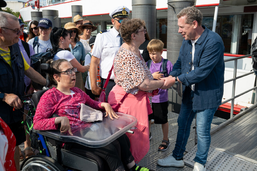 Eine Aufnahme auf einem Schiffsanleger am Bodensee. Auf der rechten Seite steht AK-Pr&auml;sident Bernhard Heinzle in Jeans und einem offenen, blauen Hemd &uuml;ber einem wei&szlig;en T-Shirt. Er l&auml;chelt und gibt einer &auml;lteren Frau im Leoparden-Print-Oberteil herzlich die Hand. Im linken Vordergrund sitzt eine junge Frau mit Brille in einem Rollstuhl mit Therapietisch und blickt zu ihnen auf. Sie tr&auml;gt ein pink gemustertes Oberteil. Ein &auml;lterer Mann steht unterst&uuml;tzend hinter ihrem Rollstuhl. Im Hintergrund wartet eine Gruppe weiterer Fahrg&auml;ste auf dem Steg, darunter ein Kapit&auml;n in wei&szlig;er Uniform und Schirmm&uuml;tze. Im rechten Hintergrund ist der Eingangsbereich eines wei&szlig;-roten Passagierschiffs zu sehen.