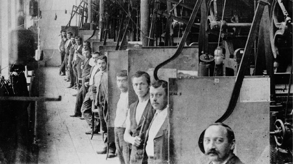 Eine historische Schwarz-Wei&szlig;-Aufnahme aus dem Inneren einer Fabrik. Eine lange Reihe von Arbeitern steht hintereinander an ihren jeweiligen Arbeitsstationen, die durch trennende Platten voneinander abgeteilt sind. Die M&auml;nner blicken alle in Richtung der Kamera. Im Vordergrund ist das Gesicht eines Mannes mit Schnurrbart angeschnitten zu sehen. Entlang der Decke und W&auml;nde verlaufen komplexe mechanische Vorrichtungen, Riemen und Maschinenteile. Der Boden besteht aus Holzdielen.