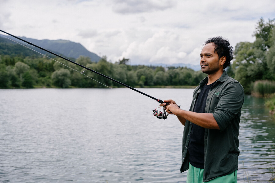 Ein Mann steht am Ufer eines ruhigen Sees und h&auml;lt eine Angelrute in den H&auml;nden. Er blickt l&auml;chelnd nach links ins Profil. Er hat dunkles, lockiges Haar und tr&auml;gt ein offenes, dunkelgr&uuml;nes Hemd &uuml;ber einem schwarzen T-Shirt sowie eine hellgr&uuml;ne Hose. Im Hintergrund erstreckt sich eine bewaldete H&uuml;gellandschaft unter einem bew&ouml;lkten Himmel.